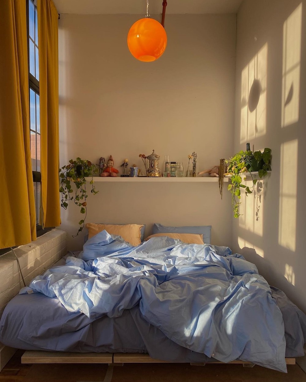 a small bedroom with pastel blue bedding, an open shelf above the bed, and real houseplants