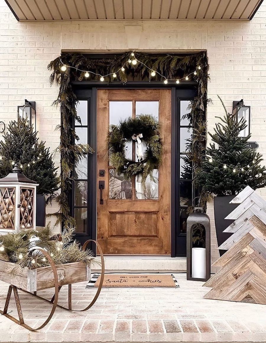 a front porch with outdoor christmas decorations including a sleigh, a wooden tree, real pine trees, garland, wreaths, and lights