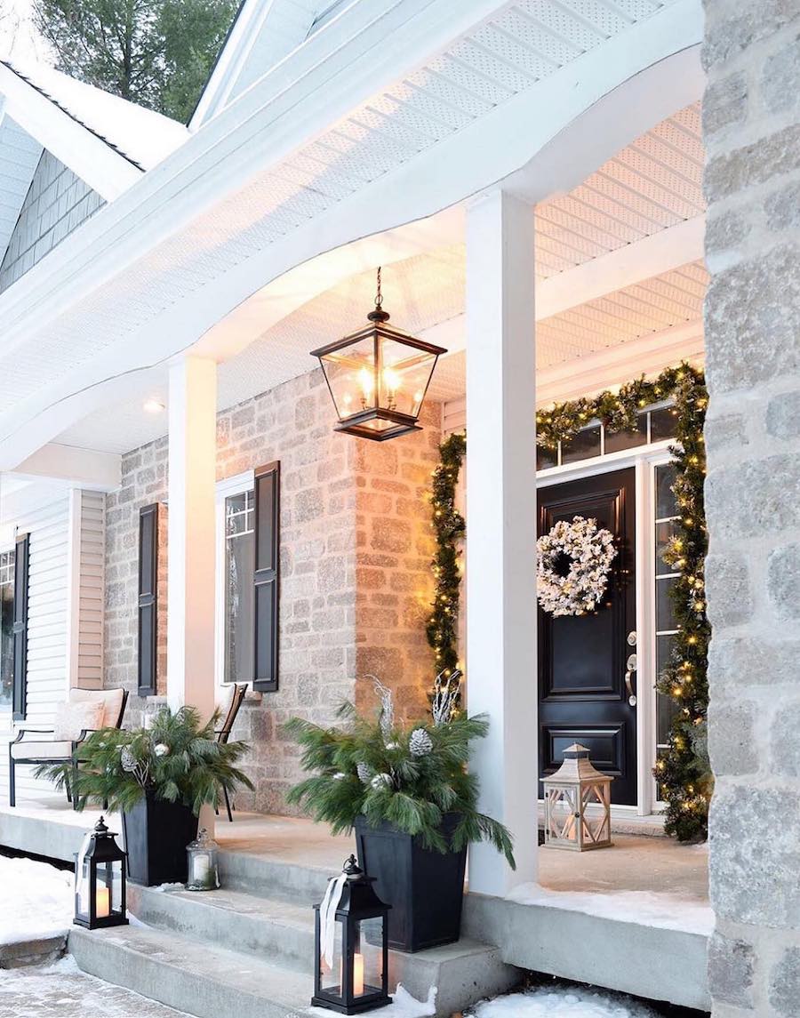 a front porch with lanterns, pine garland, a snowy wreath, and small pine trees