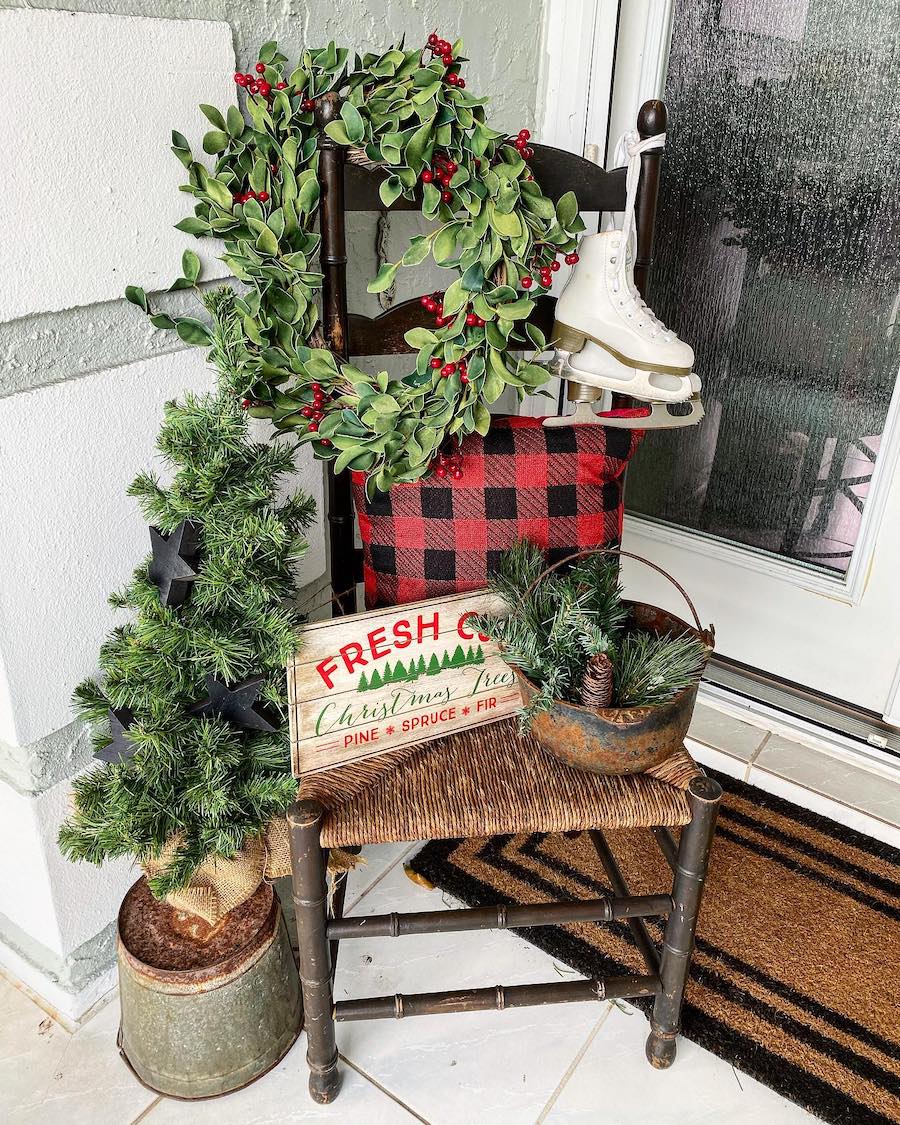 a front porch with outdoor christmas decor including a wooden chair with a holly berry wreath, a small pine tree, a plaid pillow, and a christmas sign