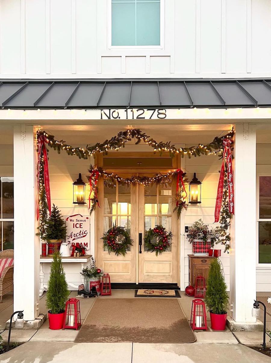 a front porch decorated with small pine trees, lights, red garland, and wreaths