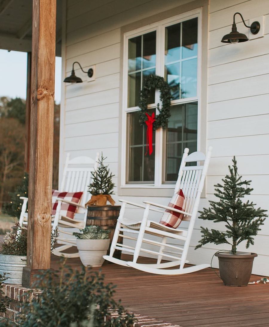 a front porch with white rocking chairs, plaid pillows, wreaths, and small pine trees