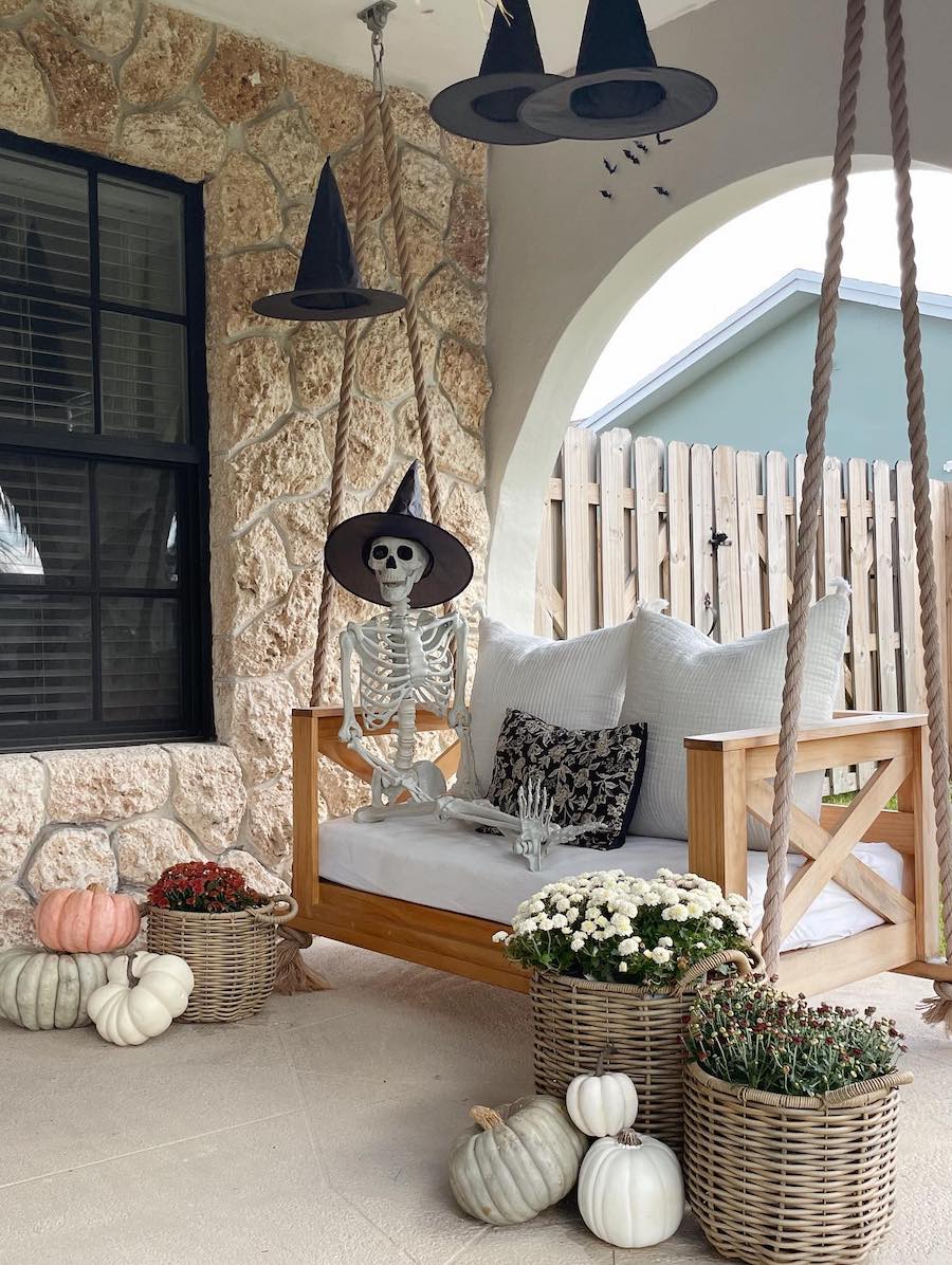 a front porch featuring a swing with a skeleton wearing a witch hat sitting on it, pumpkins, florals, and more floating witch hats