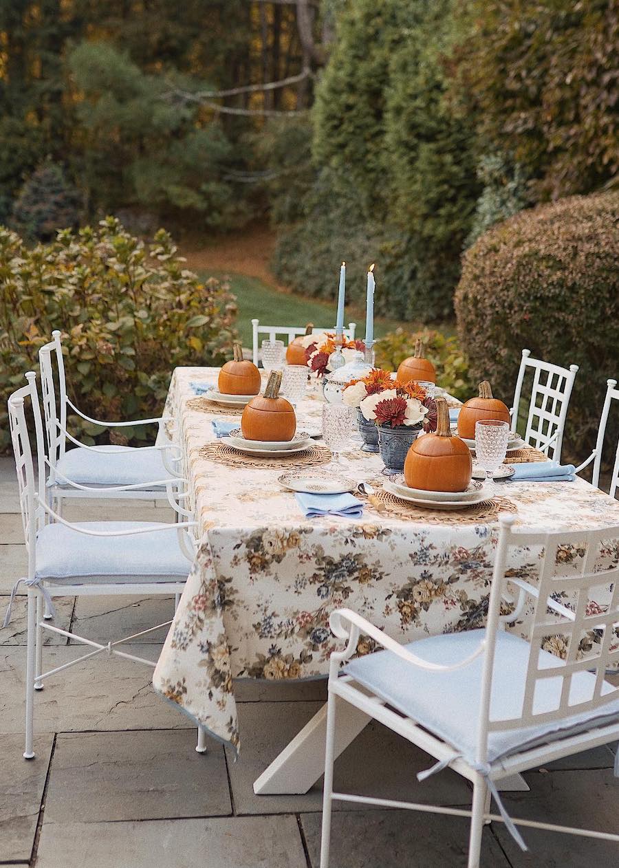 a table outside with outdoor fall decor ideas including pumpkin bowls, fall florals, a vintage floral table cover, and candlesticks